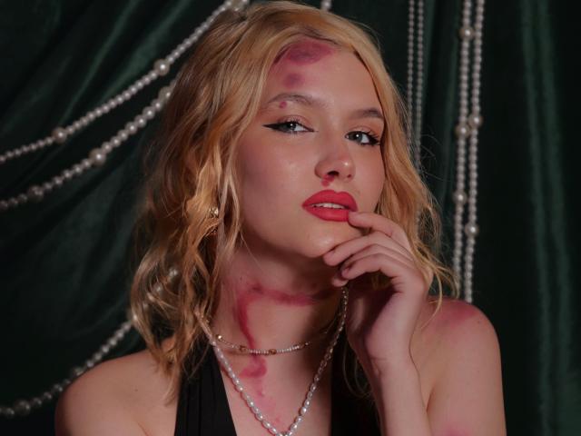 VelvetSkyy with blonde wavy hair, pink makeup smears on face, black halter top, pearl necklace, finger touching red lipstick, dark green backdrop, pearl beads in background.