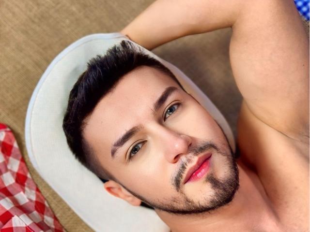 DaltonWhite, shirtless, with short dark hair, green eyes, and a trimmed beard, lying on a white hat, right arm raised, looking at camera, red plaid blanket partially visible.