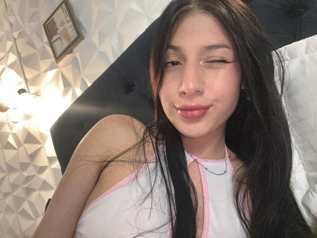 SophieBeaumont with long black hair, winking, wearing a pink-trimmed white halter top, smiling with pink lipstick, sitting on a dark headboard, light glowing from the left.