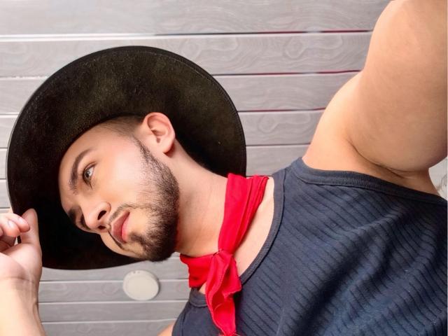 DaltonWhite, con un sombrero negro, una bandana roja y una camisa oscura con textura, mostrando un brazo musculoso y una barba recortada, sosteniendo el borde del sombrero. Su rostro está inclinado hacia un lado..
