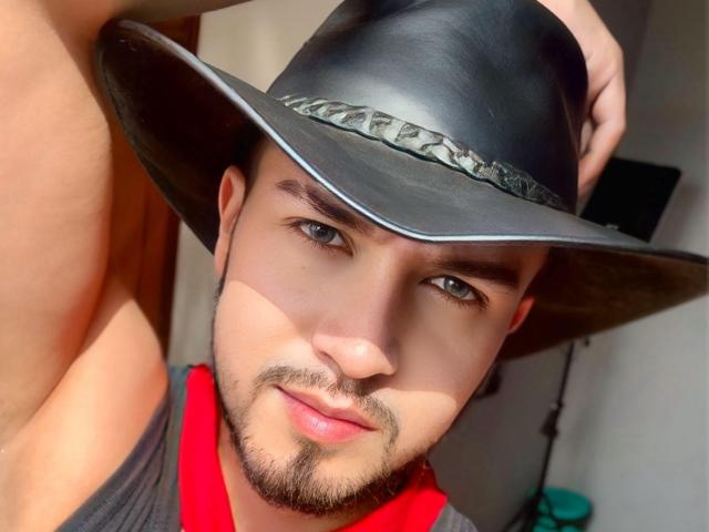 DaltonWhite with a black leather cowboy hat, sporting a red shirt, light brown skin, blue eyes, and a beard, looking at the camera with a slight smile.