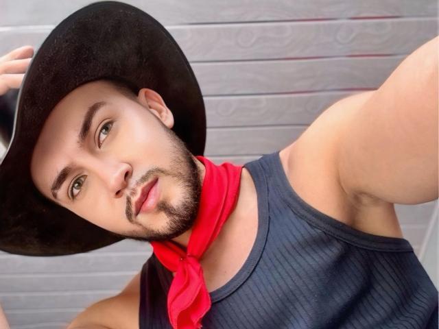 DaltonWhite heeft een zwarte cowboyhoed, een rode bandana en een mouwloos donker shirt, waardoor een gespierde arm en een getrimde baard zichtbaar zijn. Zijn groene ogen en lichte glimlach zijn te zien..