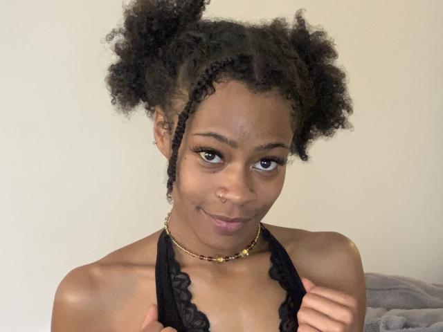 DeviantDynasty ha i capelli ricci e scuri raccolti in chignon voluminosi, indossa un top nero in pizzo con spalline sottili, sorride mostrando un piercing al naso e una collana di perline, tenendo le spalline del top..
