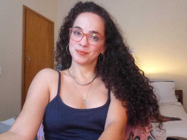 AmyFlores, met krullend zwart haar, rode bril en een zwart tanktopje, glimlacht naar de camera en laat haar kleine borsten en een zilveren ketting zien. De achtergrond is een slaapkamer..