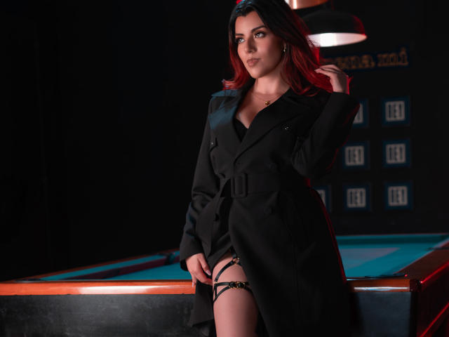 ZoeeMar with red hair, black coat, and black lingerie, standing by a pool table, showing her glistening pussy through the coat's hem, looking confident in a dimly lit room.