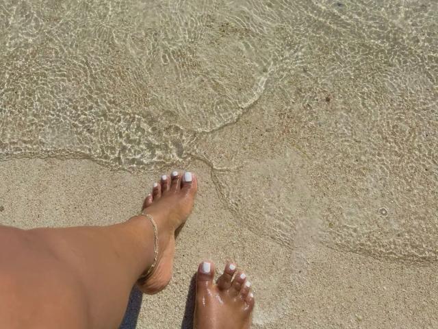 KatyBomb's tan feet with white-painted toes and a silver anklet are in shallow, clear water, showing wet sand and small ripples.