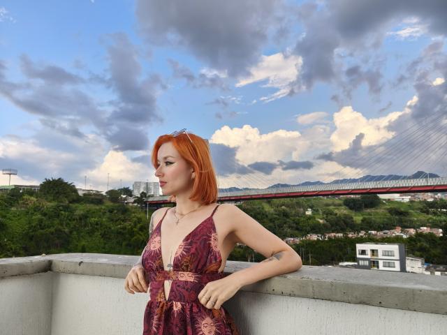SarahRousen with red hair, wearing a deep V, floral dress, showing her small tits and tattooed arm, leaning on a concrete ledge, against a cloudy sky.