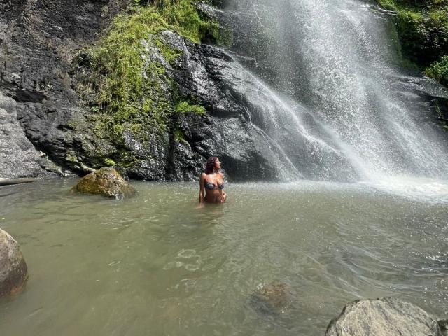 AmberQueeny with a muscular, shirtless body stands in a waterfall pool, water up to her ass, dark wet hair, arms crossed, rocky waterfall in the background.