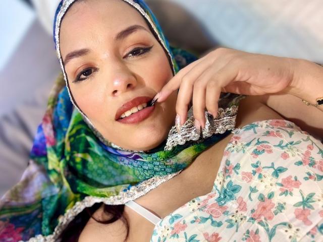 AliceKayleigh with her headscarf, floral dress, and lace trim, holding her lip with a finger, showing her tits and cute smile. She's got sexy brown eyes and pink lips.