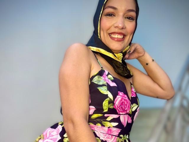 AliceKayleigh with dark brown hijab and yellow trim, biting her finger, wearing floral black dress, showing her tits, light skin, pink lipstick, and black nail polish.