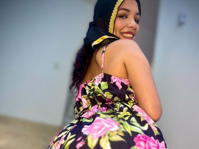 AliceKayleigh with a smile, wearing a floral dress, black headscarf with yellow trim, and visible tits, posing with one hand near her face, showing a cute, confident look.