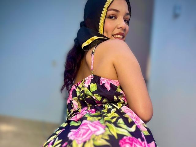 AliceKayleigh with dark hair, wearing a floral dress, black headscarf with yellow trim, smiling, showing her back and ass, pink roses on black fabric, looking over her shoulder.