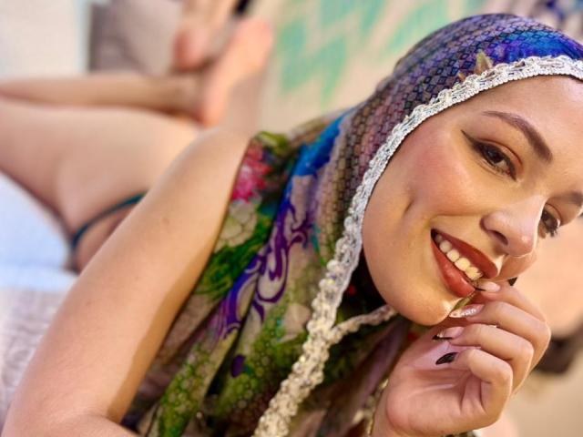 AliceKayleigh with a green and purple headscarf, lying on her stomach, wearing green lingerie and white lace panties, showing her ass and red lipstick, looking at the camera.