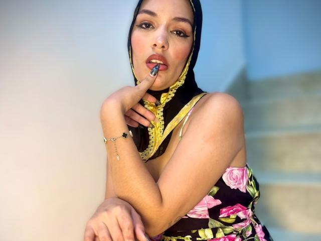 AliceKayleigh with black and yellow headscarf, smiling, showing off her tits in a floral dress, sitting with arms crossed, blue wall in background, long nails, bracelet.