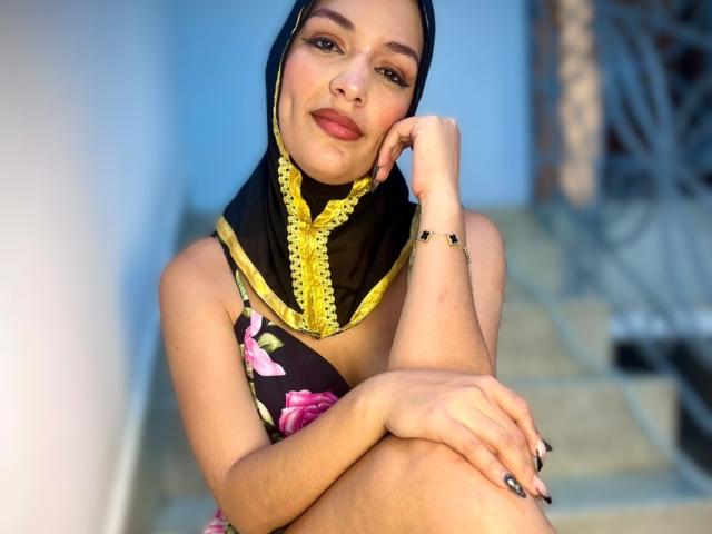 AliceKayleigh with a black and yellow headscarf, floral dress, and finger in mouth, showing her tits and holding a lipstick. She's on stairs, looking seductive.
