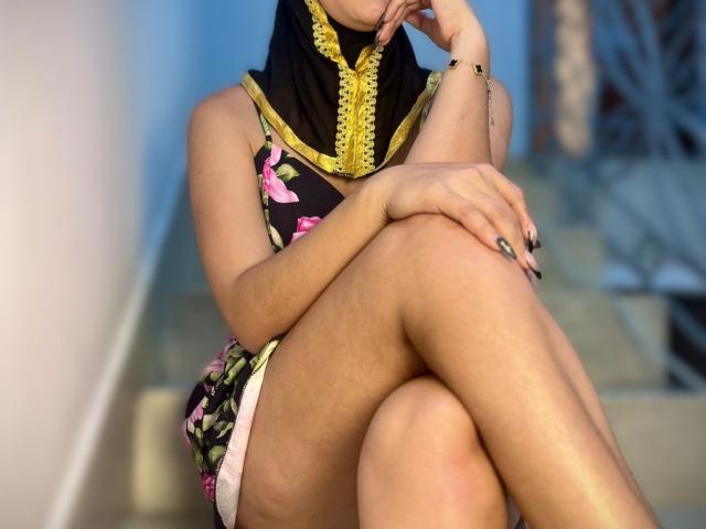 AliceKayleigh with a black and yellow headscarf, pink and green floral dress, smiling, hand near mouth, sitting, visible boobs, medium skin, dark nails, gold bracelet, looking up.