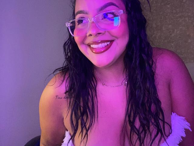 AmarantaCarey with curly black hair, purple-tinted glasses, and white frilled top, smiling at the camera, showing her tits, with a tattoo on her right shoulder.