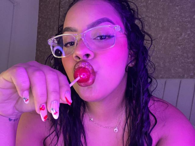 AmarantaCarey with wet black hair, purple-tinged skin, clear glasses, and red lipstick, sucking a pink lollipop, showing red and white painted nails, and a silver necklace.