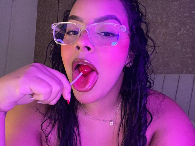 AmarantaCarey with wet black hair, clear glasses, and red lipstick, sucking on a red lollipop, holding it near her glossy, pink-lit lips, and showing her red nails.