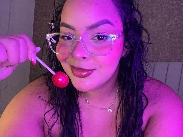 AmarantaCarey with wet black hair, wearing clear glasses, holding a red lollipop near her glossy pink lips, smiling, and showing a tattoo on her right shoulder.