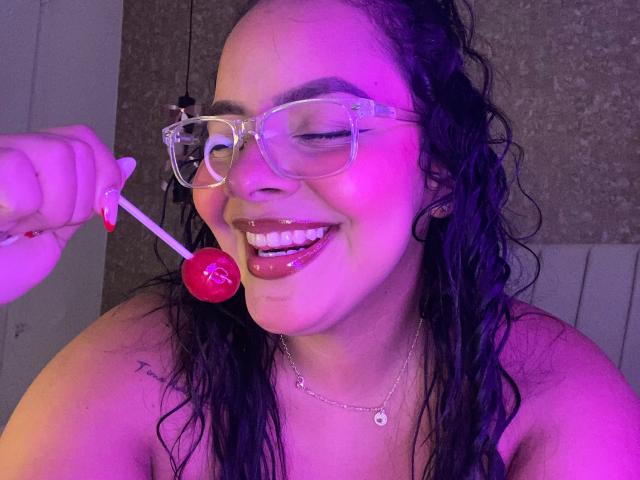 AmarantaCarey with wet black hair, clear glasses, and glossy pink lips, smiling while holding a red lollipop to her mouth. Purple lighting highlights her shiny face and chest.