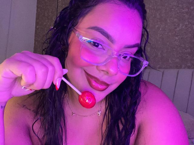 AmarantaCarey with wet black hair, pink glasses, and red lipstick, holding a red lollipop near her glossy pink lips, smiling. Her tits are partially visible. Purple light.