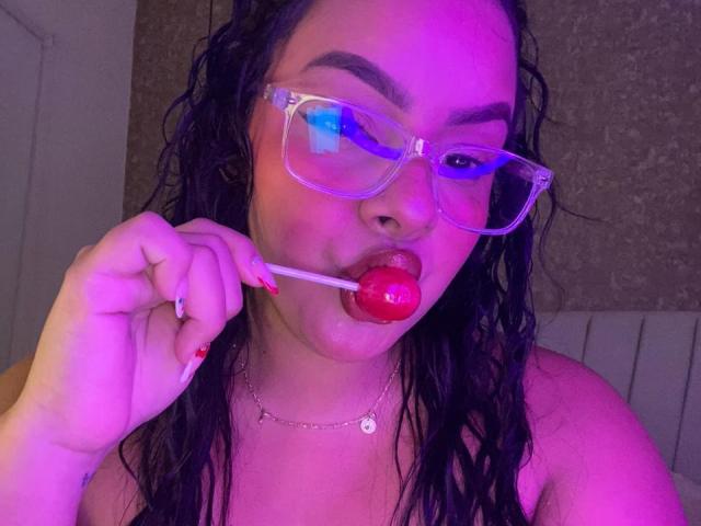 AmarantaCarey with wet black hair, wearing transparent glasses, sucking a red lollipop with her pink-manicured hand, purple and blue lighting on her face, necklace with small pendant.