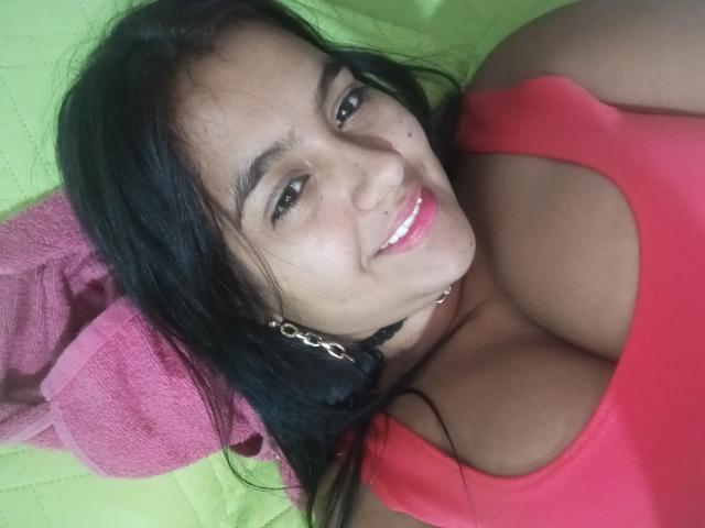 AmberMedellin a les cheveux foncés, les yeux marron, et elle sourit à l'appareil photo. Elle porte un débardeur rouge qui met en valeur sa poitrine généreuse, avec une serviette rose et un fond vert..