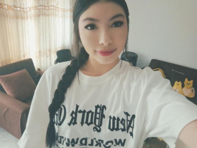 ViolettaLeroy with long black braid, wearing a white "Dark Amsta" t-shirt, soft smile, light skin, cute, small nose, dark eyes, standing in a bright room.