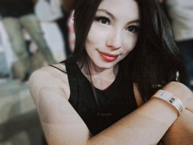 ViolettaLeroy with long dark hair, smiling, wearing a black sleeveless top, arms crossed, red lipstick, and a white wristband, in a blurry indoor setting.