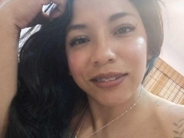 Paulina Colemann a les cheveux foncés, un trait d'eye-liner prononcé et un collier en argent. Elle sourit, on voit des bagues dentaires sur ses dents, sa main est dans ses cheveux, et elle montre un tatouage sur son épaule..