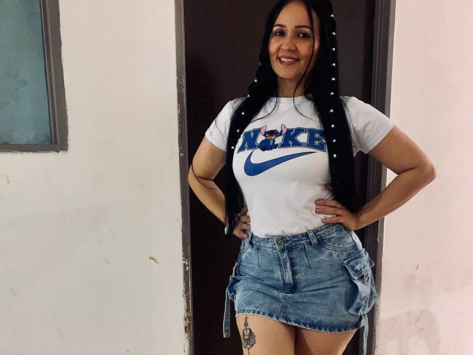 Karus with long black braids, white Nike shirt, blue denim skirt, hands on hips, smiling, visible tattoo on right thigh, standing in doorway. Boobs, ass, tits, pussy, hot.