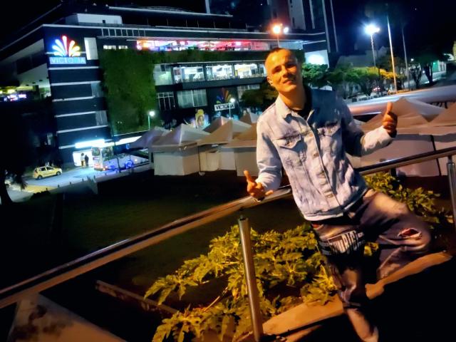 StrongAlan, wearing a light blue denim jacket and ripped jeans, stands on a metal railing at night, thumbs up, with a confident smile, near a brightly lit building.