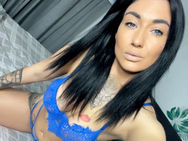 BabyMarie, com cabelo preto, usando um sutiã de renda azul e com tatuagens, sentada em uma cama, mostrando os seios e uma tatuagem nos lábios, olhando para a câmera com uma expressão de desdém..