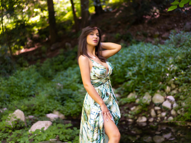 HarperSkye with long brown hair, wearing a green patterned dress, standing in a forest, hand in hair, showing her tits and a sexy leg up. Her eyes are closed.