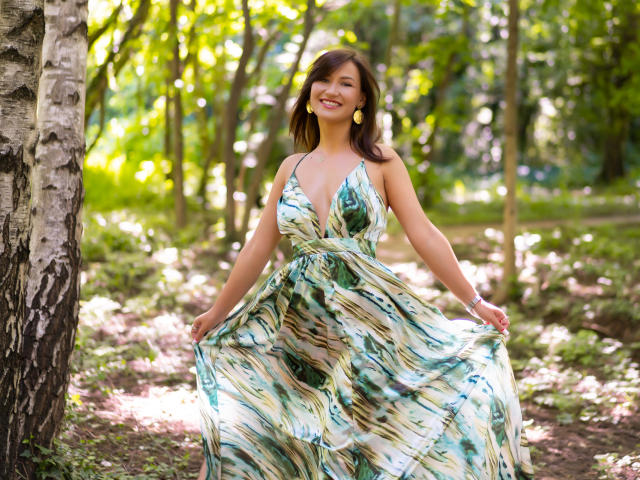 HarperSkye with medium tits, smiling, wearing a green and white patterned dress, holding it out, yellow earrings, brown hair, standing by a tree in a sunny forest.