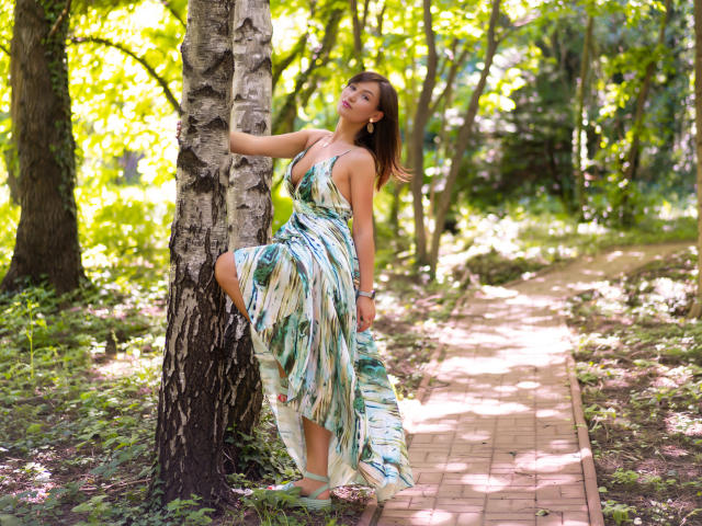 HarperSkye is standing against a birch tree, wearing a green and white patterned dress, with one leg raised, showing her tits, in a sunny forest path.