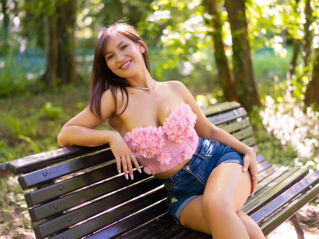HarperSkye with pink flowered strapless top, showing her tits, and denim shorts, sitting on a wooden bench in a sunny forest, smiling, arms resting.