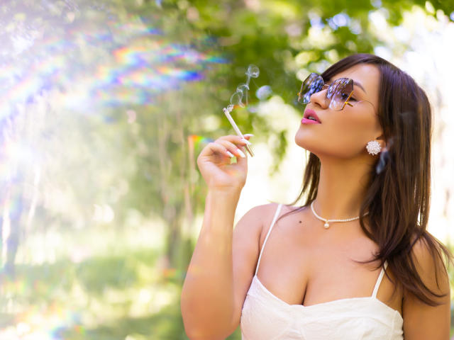 HarperSkye with tits visible in a white dress, holding a cigarette, blowing bubbles, wearing sunglasses, and a flower earring, standing outside in sunlight.