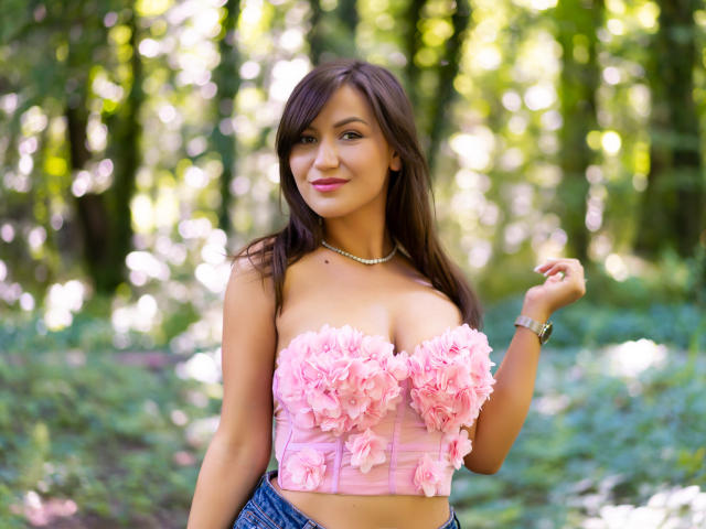 HarperSkye with pink flowered tube top, showing her tits, wearing a necklace, and blue jeans, standing in a forest, smiling, with long brown hair and a watch.