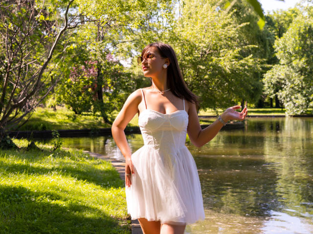 HarperSkye stands in a sunlit, grassy area, wearing a white, sleeveless, strapless dress that shows her tits, with a necklace, earrings, and a calm, confident expression.