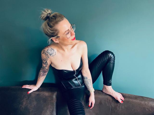 MiaHase-hot with blonde bun, glasses, red lipstick, black leather bustier, and tight black pants, tattooed arms, sitting on brown couch, blue wall, red nails, looking to the side.
