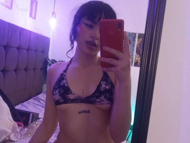 YessBoobs with dark hair, wearing a black lace bra, holding a red phone, has a tattoo below her tits, mirror selfie, purple light in bedroom.