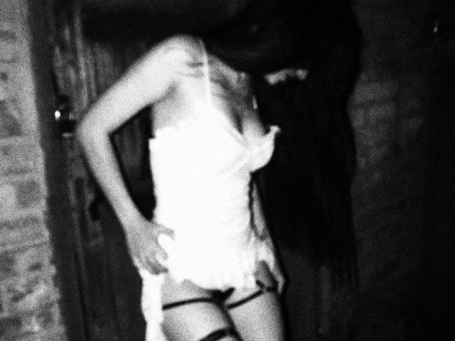 AriXoxo mit langen, dunklen Haaren, trägt ein weißes Spitzen-Babydoll und ein schwarzes Strumpfband, ihre Brüste sind leicht sichtbar, sie steht mit den Händen auf ihrem Hintern, auf einem Schwarz-Weiß-Foto..