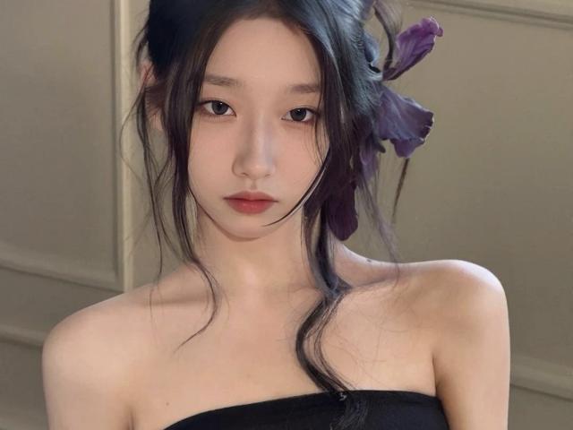 LinlinYa, con i capelli scuri, un fiore viola e le spalle scoperte, indossa un top nero senza spalline, guarda leggermente verso il basso con un'espressione neutra, con una luce soffusa e una pelle chiara..
