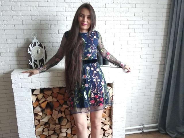 GinaSoull with long brown hair, wearing a floral navy dress, standing in front of a white brick fireplace, arms resting on the mantle, showing off her tits.