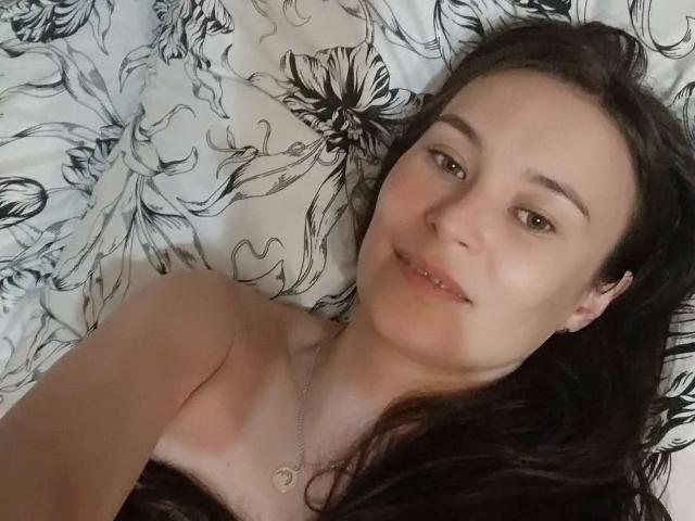 GinaSoull with dark hair lying on a bed, smiling at the camera, showing bare shoulders and wearing a necklace with a heart pendant, looking cute and relaxed.
