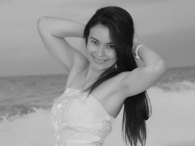 Black and white photo of GinaSoull. She's a young woman with long, straight dark hair, smiling at the camera. She's wearing a strapless lace dress, with both arms raised, hands in her hair. She's got a bracelet on her right wrist. Ocean and waves in the background, slightly blurred. GinaSoull has a confident, relaxed expression. Her skin is light, and she's looking directly at the camera. The texture of the lace dress is detailed, contrasting with the smoothness of her skin.