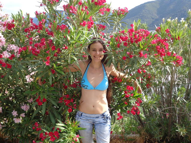 GinaSoull with small tits, blue bikini top, and blue jeans, standing in front of red and pink flowers, smiling, arms out, wearing a brown leather jacket.