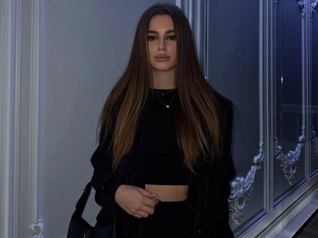 StellaHoneyX has long brown hair, wearing a black crop top, black jacket, and black pants, with a black bag, standing near a window at night.